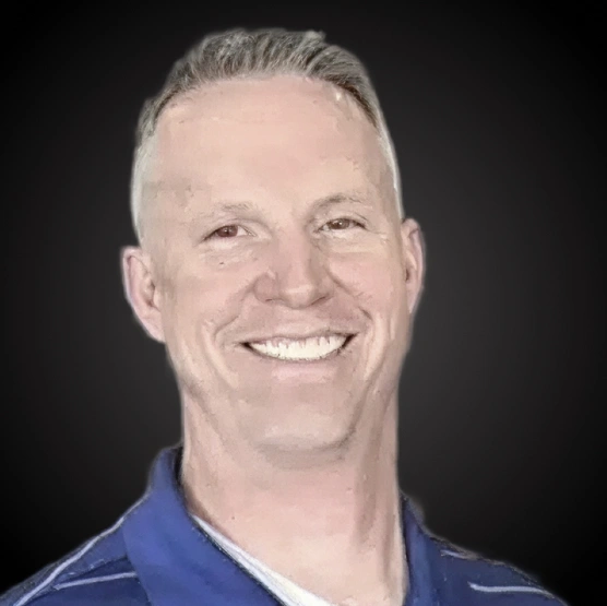 Professional headshot of male dentist smiling in blue clinical shirt against dark background.