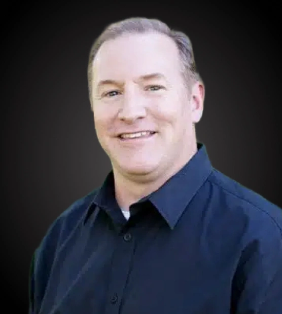 Professional headshot of male dentist smiling against dark background wearing navy blue shirt.