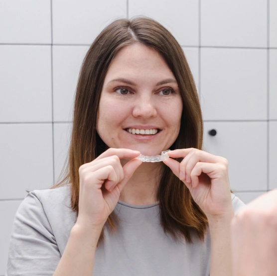 Young woman holding clear aligner in front of white tiled bathroom wall, smiling.