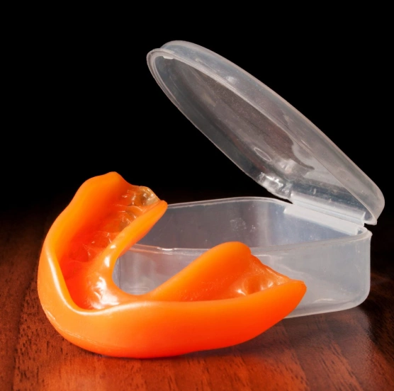 Orange mouthguard in clear plastic storage case on wooden surface against black background.
