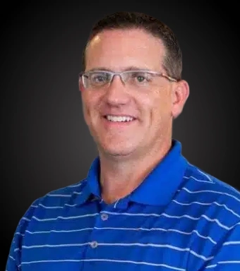 Professional headshot of male dentist wearing blue striped polo shirt and glasses against dark background.