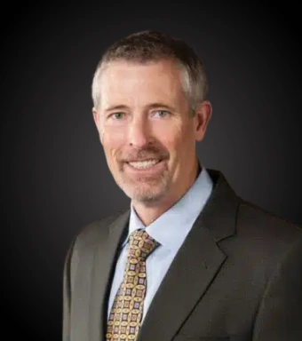 Professional headshot of male dentist in dark suit and blue shirt against dark background.