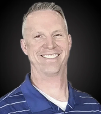 Professional headshot of smiling male dentist in blue shirt against dark background.