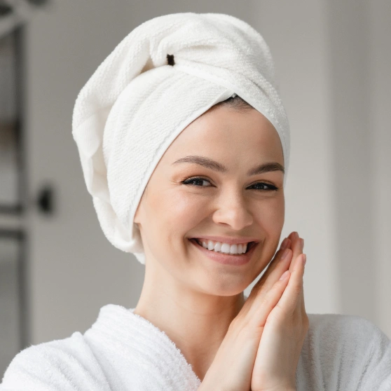 Smiling woman with towel wrapped around head touching face in bathroom setting.