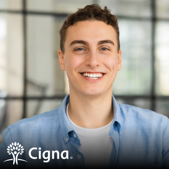 Professional headshot of smiling young man in light blue shirt, dental office setting.
